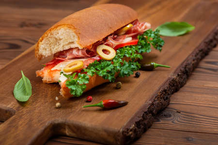 Bitten off sandwich with vegetables, spices and ham decorated greens on dark cutting wooden deskの写真素材