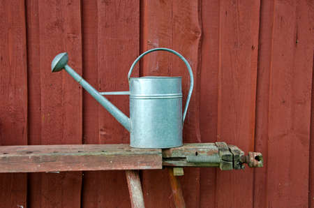 Metal watering can on a bench besides red wooden farm house wallの写真素材