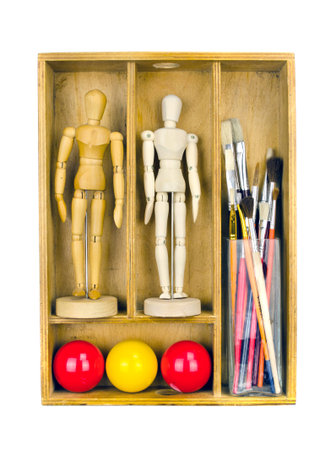 Partitioned wooden shelf filled with objects for drawing and painting - two wooden figure models, multicolored spheres and glass vase full of brushes isolated on whiteの写真素材