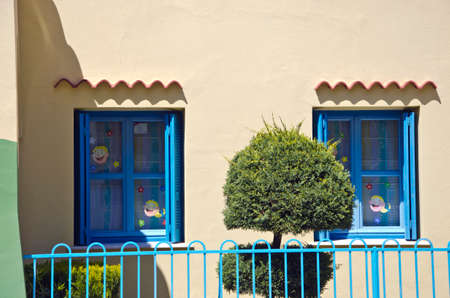Two blue windows in old yellow house decorated with children face stickers by the metal blue fence and ornamental plantの写真素材