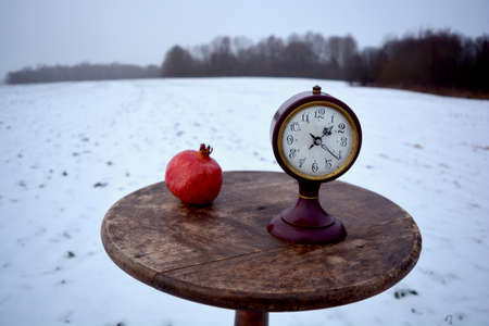 Winter still life with pomegranate and ancient clock on circle wooden retro table on snowy fieldの写真素材