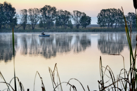 A fisherman in a boat floating on the water in the dawn sun in the fogの写真素材