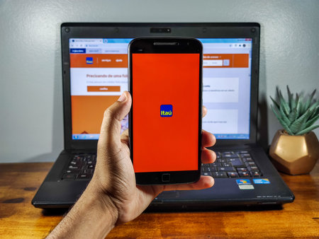 man holding smartphone with Banco ItaÃº (ItaÃº Bank) app on screen, with computer in the...