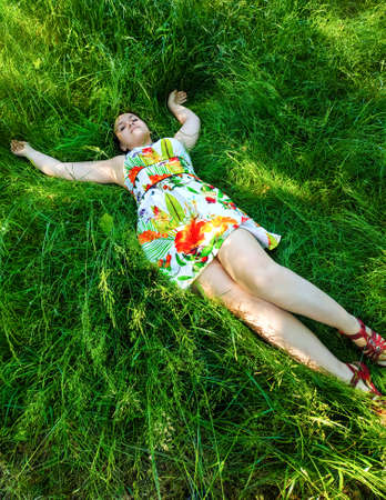 Caucasian brunette girl in a floral sundress looks at you lying among the thick green grassの写真素材