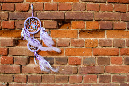 lilac-colored dreamcatcher with feathers dangling in the wind is hanging on the street with a backdrop of an old brick wallの写真素材