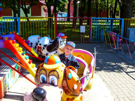 little white boy in a cap rides on a children's carousel in the summer in an amusement park smiling looksの写真素材