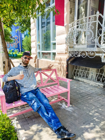 adult caucasian man with a beard is resting with a can of drink on a bench near a cafe in a small townの写真素材
