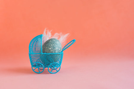 Blue Easter egg with white feathers as a baby lies in a decorative miniature blue baby carriage on a...