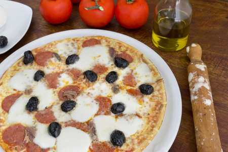 pizza with anchovies, olives, mozzarella and pepperoni surrounded by ingredients on a wooden tableの写真素材