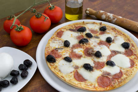 pizza with anchovies, olives, mozzarella and pepperoni surrounded by ingredients on a wooden tableの写真素材