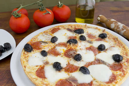 pizza with anchovies, olives, mozzarella and pepperoni surrounded by ingredients on a wooden tableの写真素材