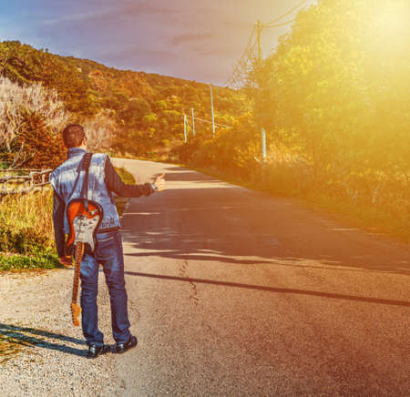 Hitchhiker with a guitar on the edge of the road at sunsetの写真素材