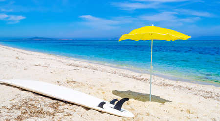 Yellow parasol and a surfboard on a white beach in the summertimeの写真素材