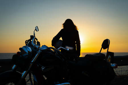 Girl and motorcycle silhouettes by the sea at sunsetの写真素材