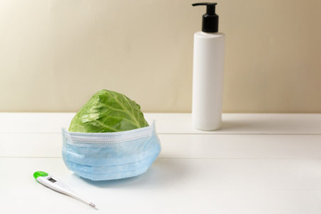 Young green cabbage in a protective mask, a sanitizer and a thermometer on a white background....