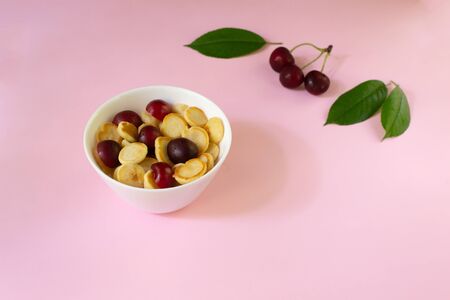 A white bowl of cereal with a tiny pancake with cherries, berries and green leaves in the background on a pink background. Copy space. Trendy food concept.の写真素材