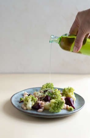 Salad of mini beets, romanesco cabbage, white beans and pouring olive oil on a gray plate on a light background. Healthy food concept. Selective focus. Vertical orientation.の写真素材