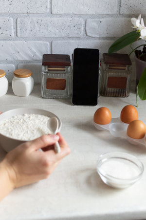 Online cooking courses. The smartphone is in the home kitchen among the products. Cooking lesson, a woman prepares a dish of flour and eggs. Copy space.の写真素材