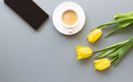 Three yellow tulips, a cup of coffee, a smartphone lies on a gray background. Spring holidays concept. Top view. Copy space.の写真素材