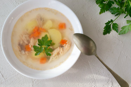 Clear fish soup with pieces of vegetables and salmon in a white bowl with a tablespoon on a white...