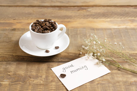 Coffee beans in a white cup with a card that reads Good morning! on a wooden background. Morning...