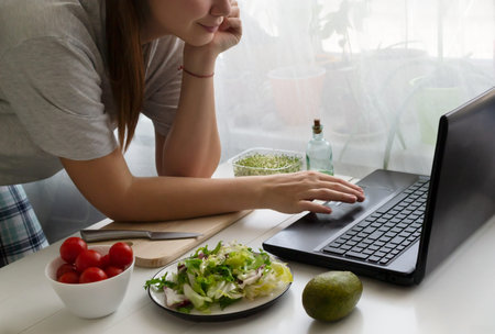 A woman is looking for a recipe for a vegan salad on her laptop. Lettuce and ingredients are on the table. Online cooking concept. Horizontal orientation.の写真素材
