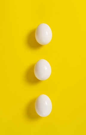 Three white chicken eggs lie on a yellow background. Vertical orientation. Top view.