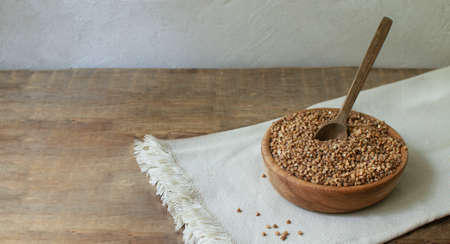Organic buckwheat groats in a wooden bowl with a spoon on a linen napkin on a wooden table. Banner. Rustic style. Healthy nutrition concept. Buckwheat contains a large amount of vitamins and minerals.の写真素材