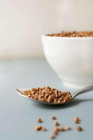A metal tablespoon with a white bowl with dry buckwheat is on a gray table. Healthy nutrition concept. Vertical orientation. Buckwheat contains a large amount of vitamins and minerals. Copy space.の写真素材