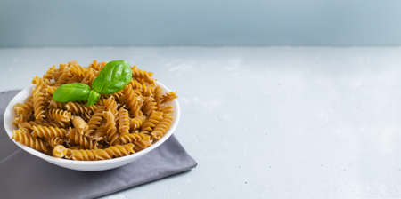 Raw fusilli in a white bowl on a gray napkin with a sprig of basal on a gray background. Time to eat pasta. Copy space. Banner. Horizontal orientation. Whole grain pasta.の写真素材