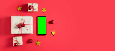 New Year's gifts are packed in craft paper and tied with red jgut and small Christmas balls on a red background. Green screen mobile phone, chroma key. Online gift shopping concept. Top view. Banner.の写真素材