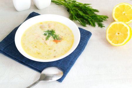 Salmon and cream fish soup in a white bowl on a blue napkin with a bunch of dill and lemon halves....