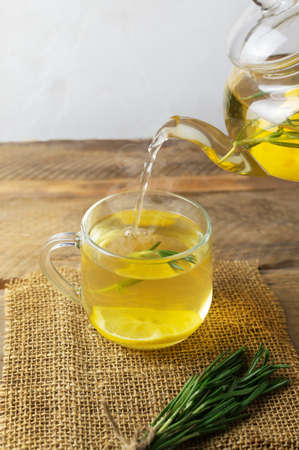 Hot warming tea is poured into a cup of rosemary, lemon and honey from a glass teapot on a wooden table. Rustic style. Alternative medicine concept. Vertical orientation. Selective focus.の写真素材