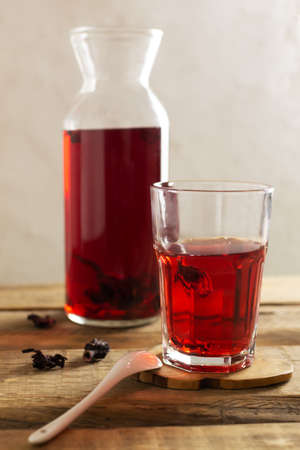 A glass cup with cold hibiscus tea stands on a stand on a wooden table. The drink has a bright red color thanks to the flowers of the Sudanese rose. Refreshing drinks concept. Vertical orientationの写真素材