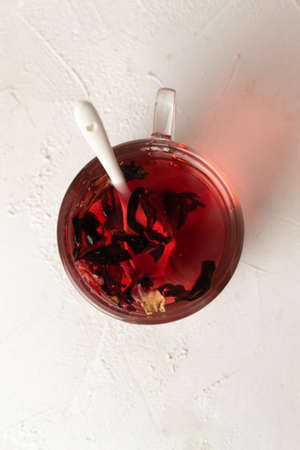 A glass cup with a teaspoon of white color with brewed hibiscus leaves on a white background. Red tea. Top view. Rosella or Sudanese rose flowers are a very popular drink in Arab countries.の写真素材