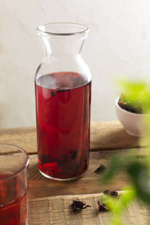 A glass jar with cold red hibiscus tea stands on a wooden table. The drink has a bright red color thanks to the flowers of the Sudanese rose. Refreshing drinks concept. Vertical orientation.の写真素材