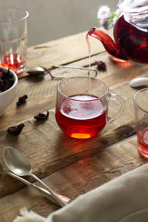 Several transparent glasses and cups sit on a wooden table. Red tea from hibiscus flowers is poured into one cup from a glass teapot. Sudanese rose flowers are a very popular drink in Arab countries.の写真素材