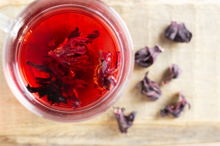 A glass cup with red hibiscus tea stands on a wooden table. The drink has a bright red color thanks to the flowers of the Sudanese rose. Alternative medicine concept. Top view. Horizontal orientationの写真素材