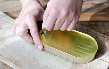A woman opens canned fish on a wooden table. Tinned fish concept. rustic style. horizontal orientation.の写真素材