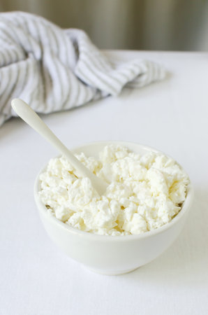 Organic cottage cheese in a white ceramic bowl with a kitchen towel on a white tablecloth. The concept of homemade food. selective focus. vertical orientation.の写真素材