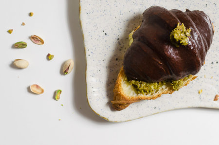 Croissant filled with pistachio paste and kadaif dough covered with dark chocolate on a white plate....