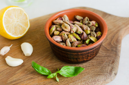 Peeled roasted pistachios in a ceramic bowl on a wooden board with basil, lemon and garlic. The process of making pistachio pesto. Horizontal orientation. Selective focus.の写真素材