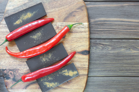 Bar of extra dark chocolate with three red chili peppers on a wooden cutting board on the table....