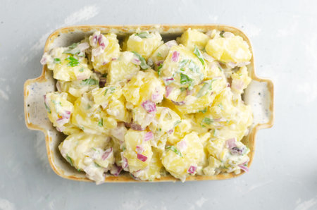 Cold potato salad with purple onion, gherkins and parsley in a white rectangular bowl on a gray table. Traditional German dish concept. Selective focus. Horizontal orientation. copy space. Top viewの写真素材