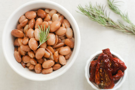 Boiled borlotti beans in a white bowl with a sprig of rosemary and sun-dried tomatoes on a gray...