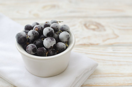 Frozen blackcurrants in a white ceramic bowl on a light background. Minimalist composition. Perfect for healthy food illustrations, vegan recipes and clean eating concepts. Copy space. Selective focusの写真素材