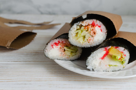 Three sushi burritos wrapped in seaweed and paper sleeves, filled with rice, seafood, lettuce, cream cheese, and tobiko. Fusion food concept combining sushi tradition with burrito style. Copy spaceの写真素材
