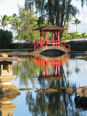 A red Japanese covered bridge crosses a calm section of pond in Hilo, Hawaiiの写真素材