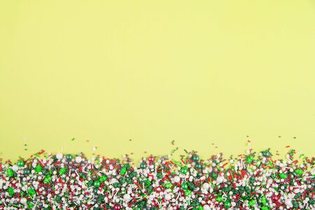 Flat lay of colorful christmas themed sprinkles along the bottom edge of a yellow backgroundの写真素材