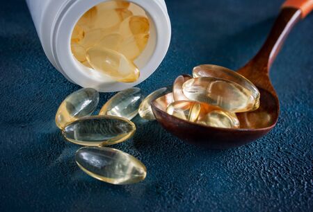 Yellow gelatin capsules with a small drug Omega-3, in a wooden spoon, on a dark blue textured background.の写真素材